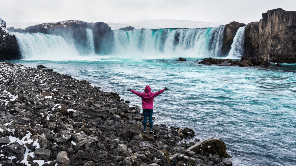 A tourist in a pink jacket standing in front of Goðafoss Waterfall in Iceland with their arms outstretched.