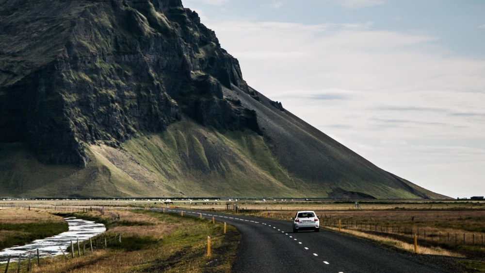 Small car driving along a road with dramatic Icelandic scenery towering overhead.